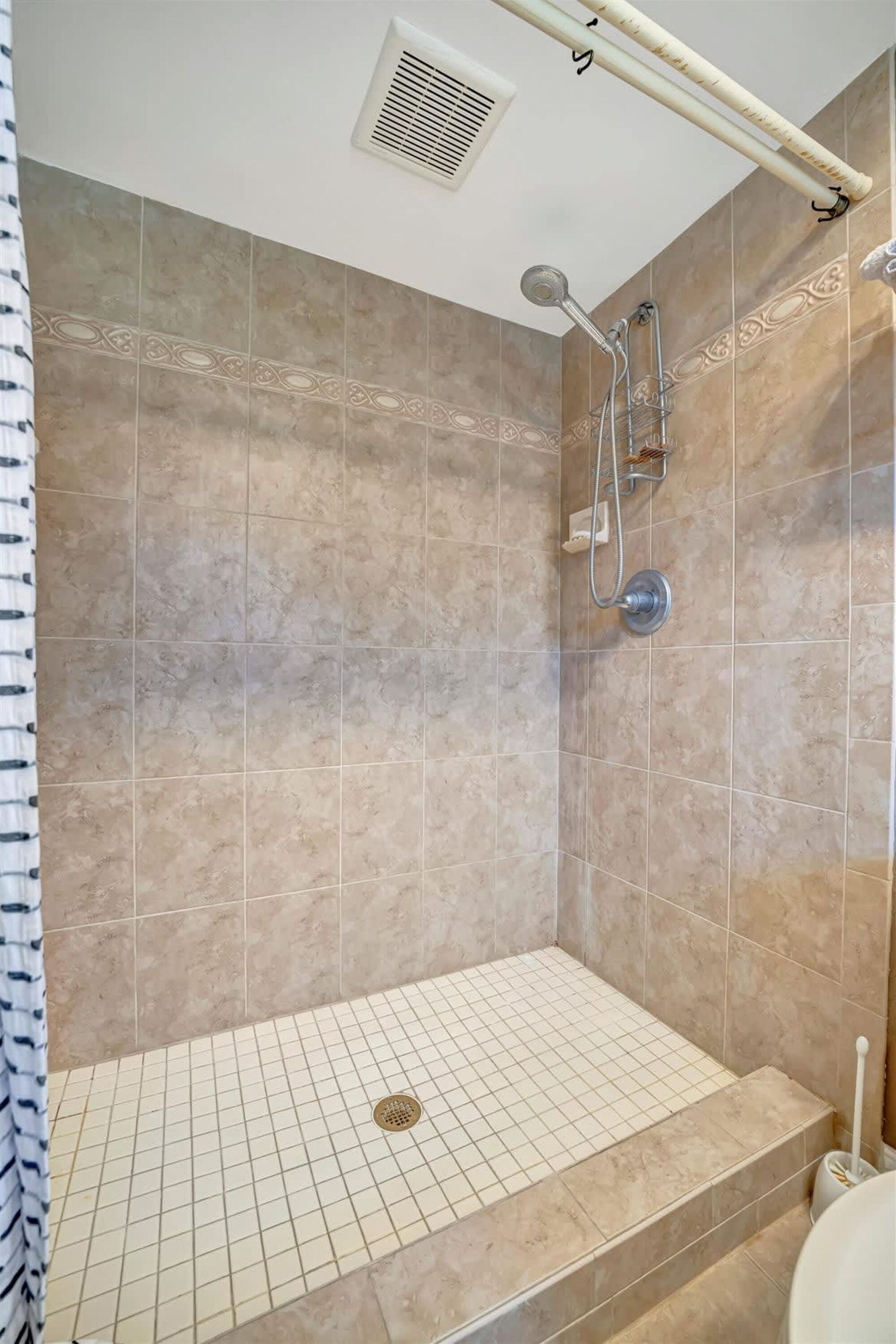 A shower area with beige tiles, a showerhead, and a handrail.