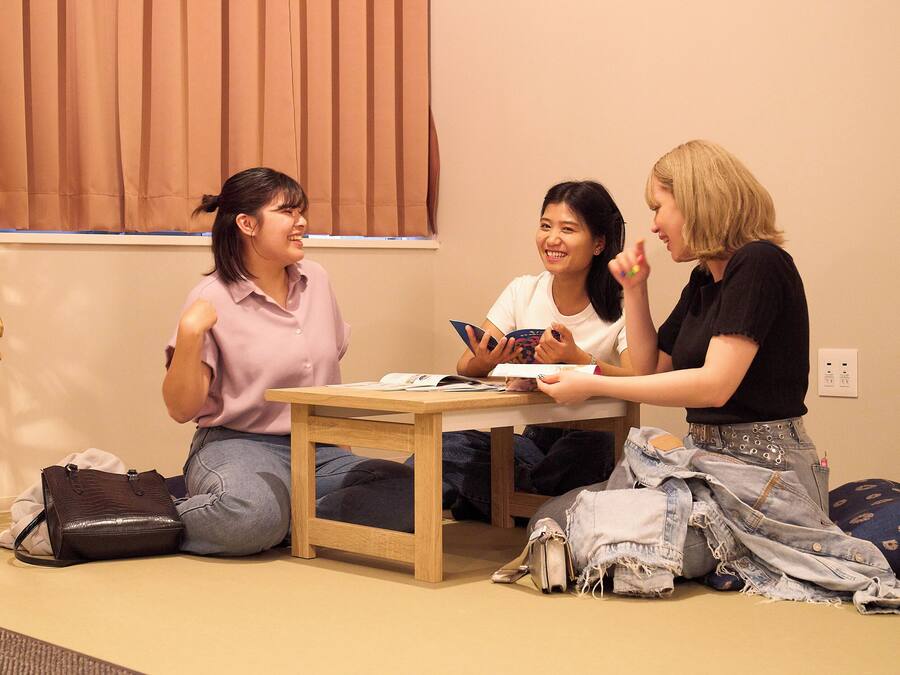 Three people sitting on the floor in a room with a table, a window with blinds, and a power outlet on the wall.