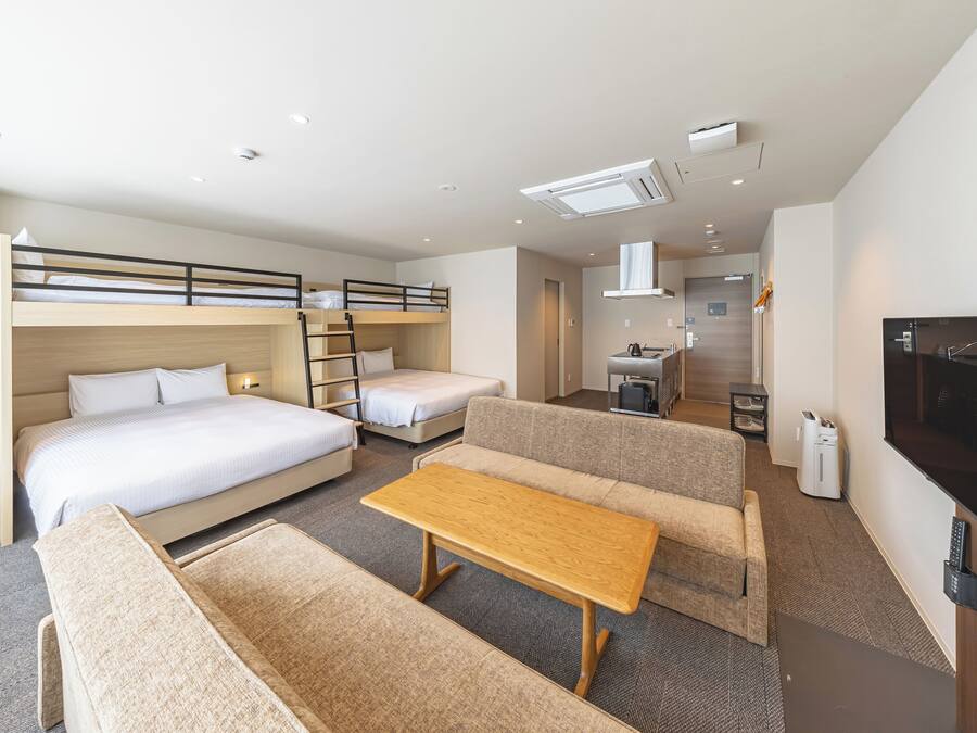 A hotel room with a bunk bed, a sofa, a table, a TV, and a kitchenette.