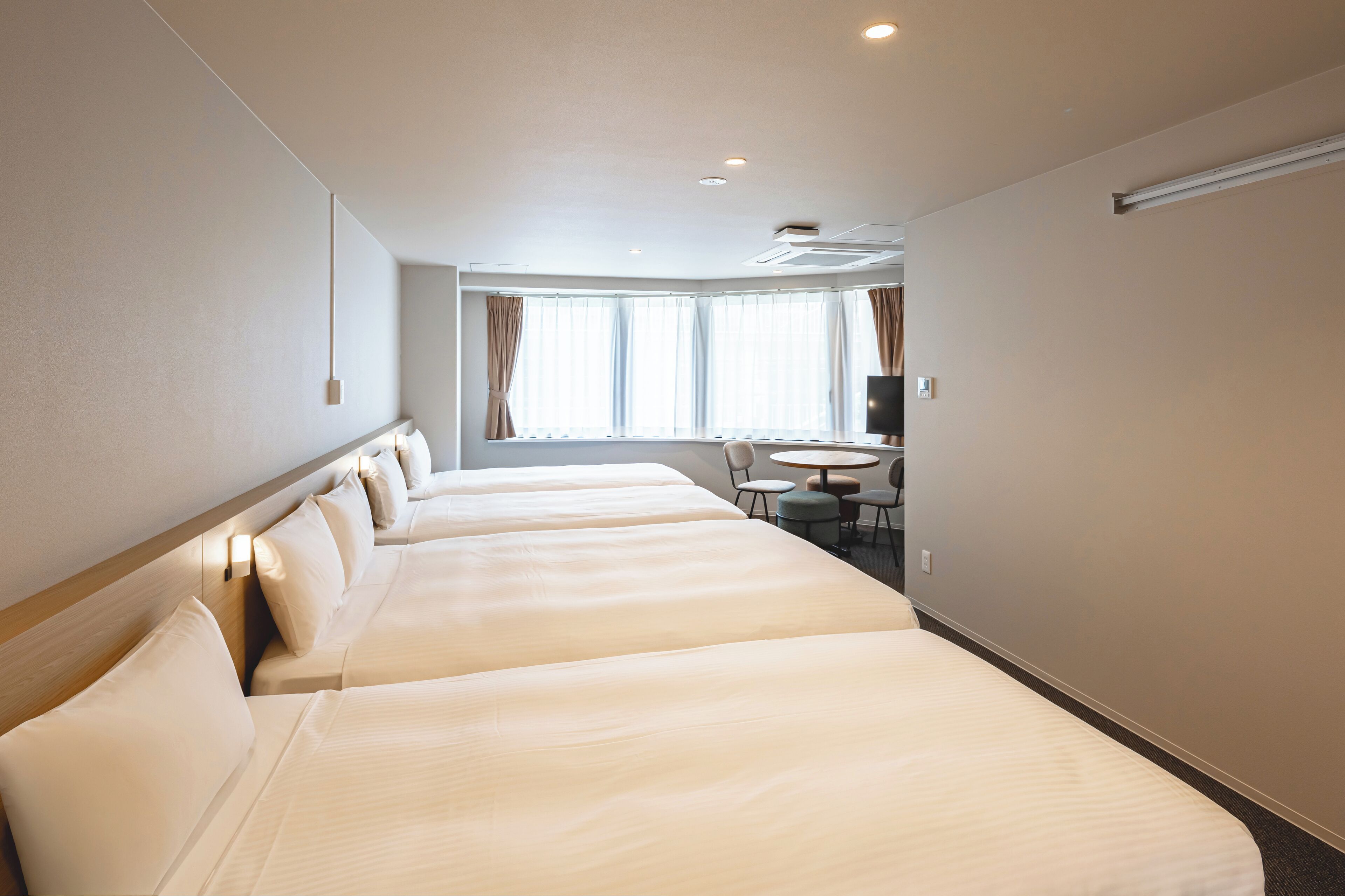 Habitación de hotel con cuatro camas alineadas, una mesita redonda y un televisor en la pared.