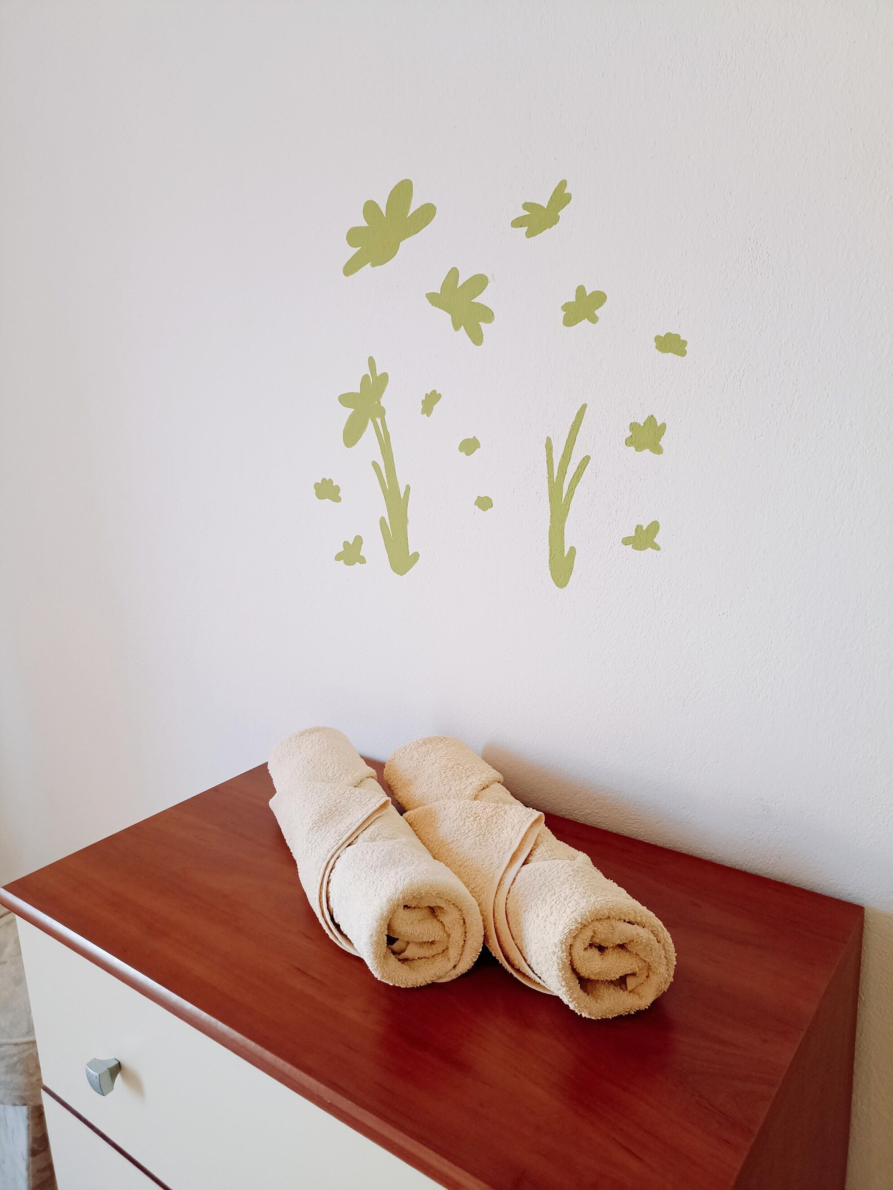 Un lit bien fait, avec deux serviettes enroulées posées sur une tête de lit en bois, un mur blanc orné d’autocollants verts et une commode blanche avec une poignée argentée.