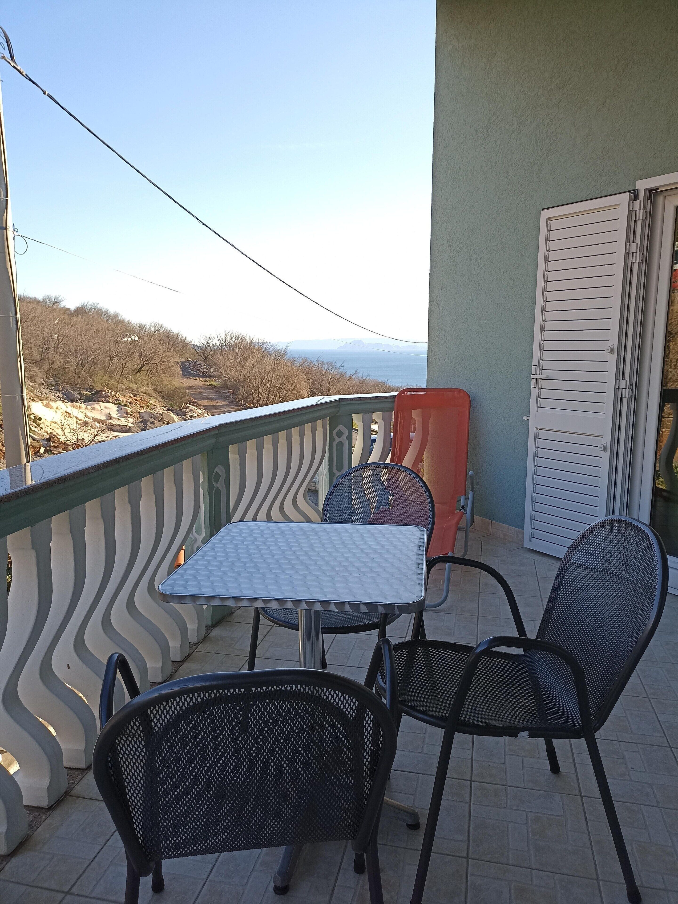A balcony with a table and chairs, a view of the sea, and a door with louvered shutters.