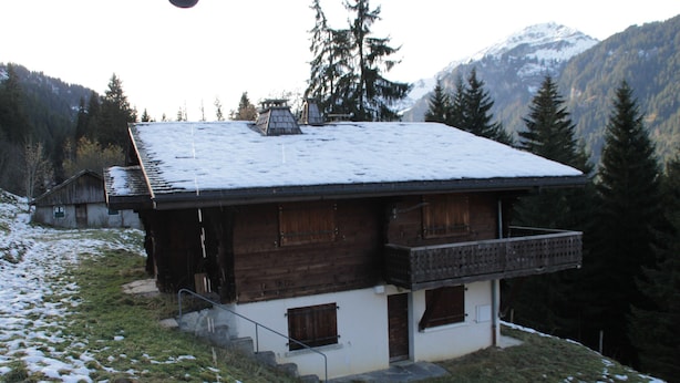 A wooden chalet with a sloped roof, a balcony, and a snow-covered landscape in the background.