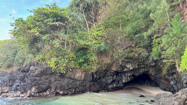 Entrada de una cueva costera con aguas turquesas cristalinas, rodeada de exuberante vegetación y terreno rocoso.