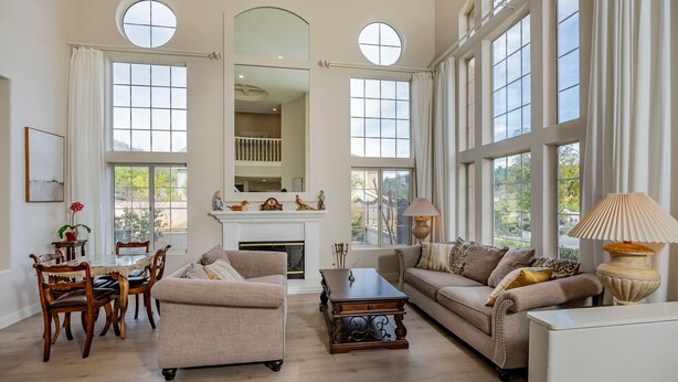 A spacious living room with high ceilings, large windows, a fireplace, and a dining area with wooden furniture.