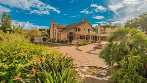 A large, single-story house with a patio area, surrounded by lush greenery and a clear blue sky.