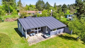 Een gelijkvloers huis met een donker dak, een overdekt terras met een tafel en stoelen, en een kleine schuur in de achtertuin.