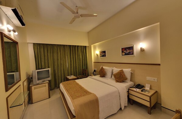 Habitación de hotel con cama, televisión, escritorio, silla, mesita de noche, teléfono y ventilador de techo.