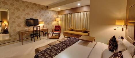 A hotel room with a bed, a desk with a chair, a sofa, a television, and a ceiling fan.