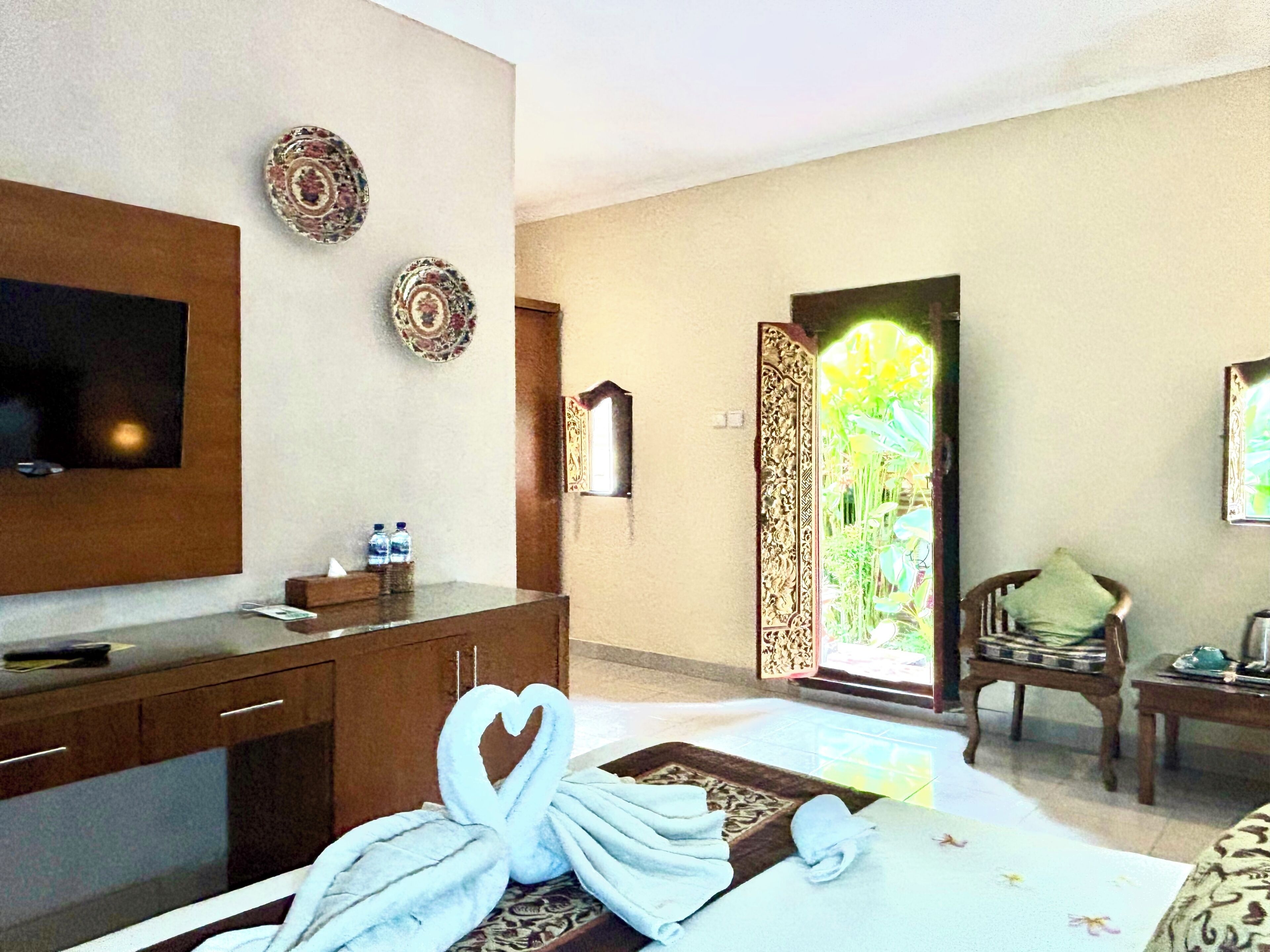 A hotel room with a bed, a TV, a desk with a chair, a decorative wall, and a door leading to a garden.