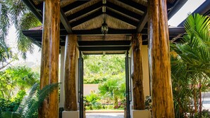 A covered walkway with wooden pillars and a tiled floor, surrounded by greenery. - Villa Essencia in Brasilito (Cabo Velas)
