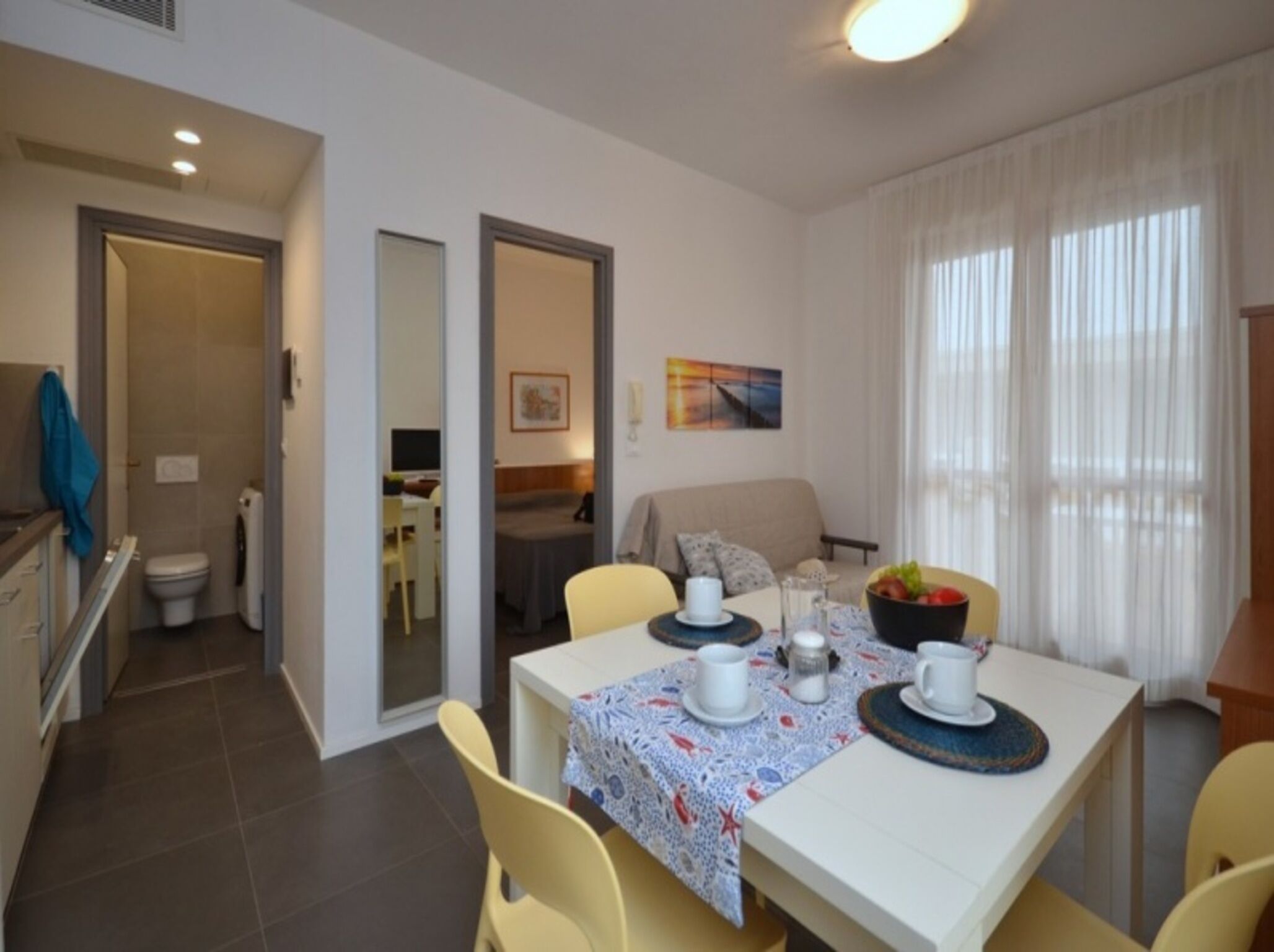 A dining area with a table set for four, a sofa, and a window with sheer curtains.