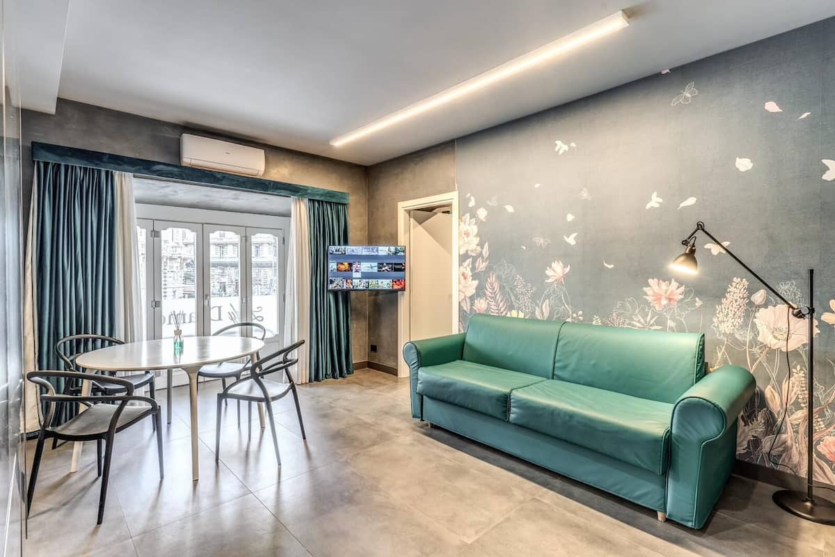 Un salon moderne avec un canapé vert, une table à manger avec des chaises, un téléviseur à écran plat et une fresque murale à motifs floraux.
