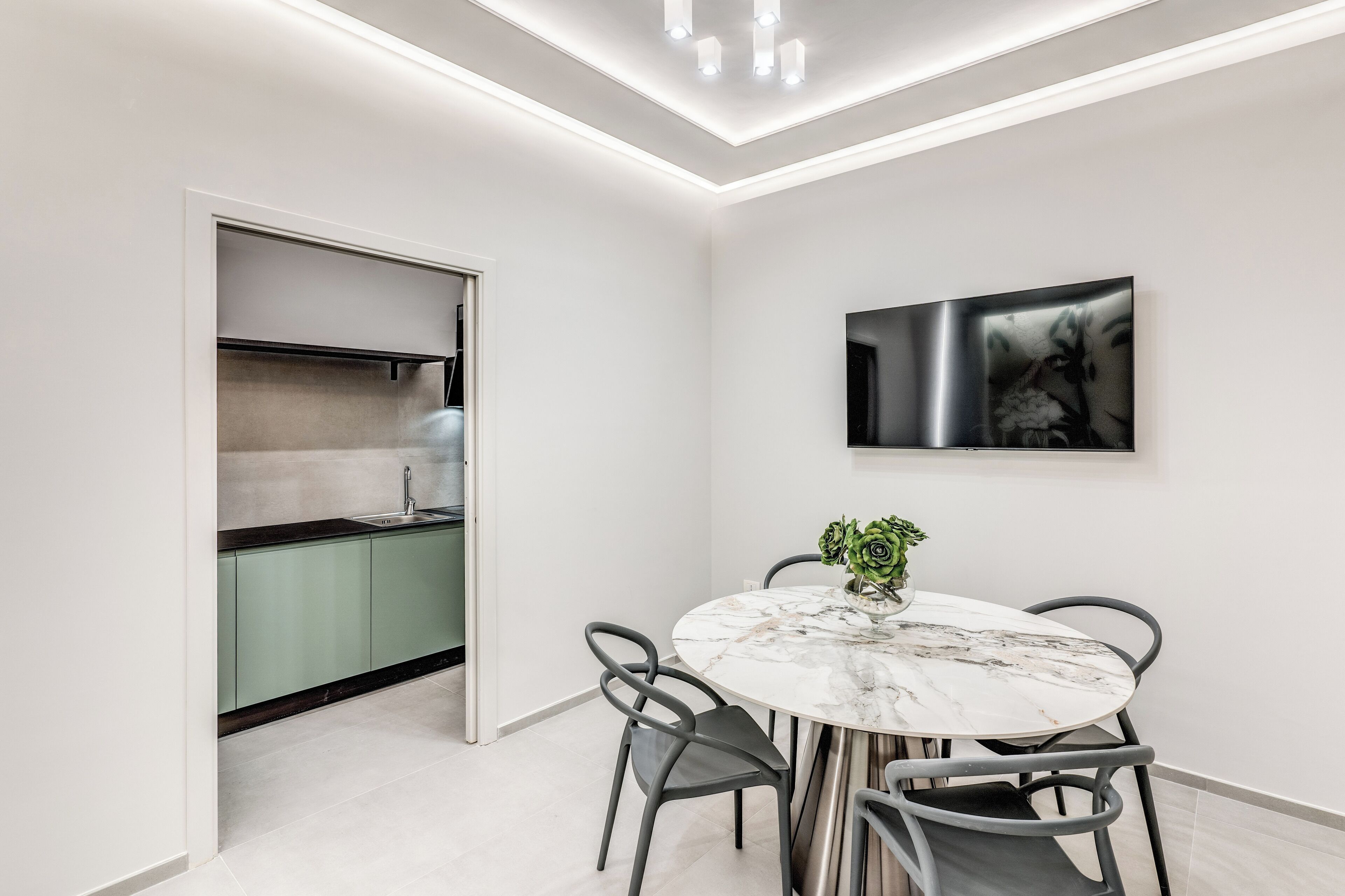 A modern kitchen with green cabinets, a marble dining table, and a flat-screen TV mounted on the wall.
