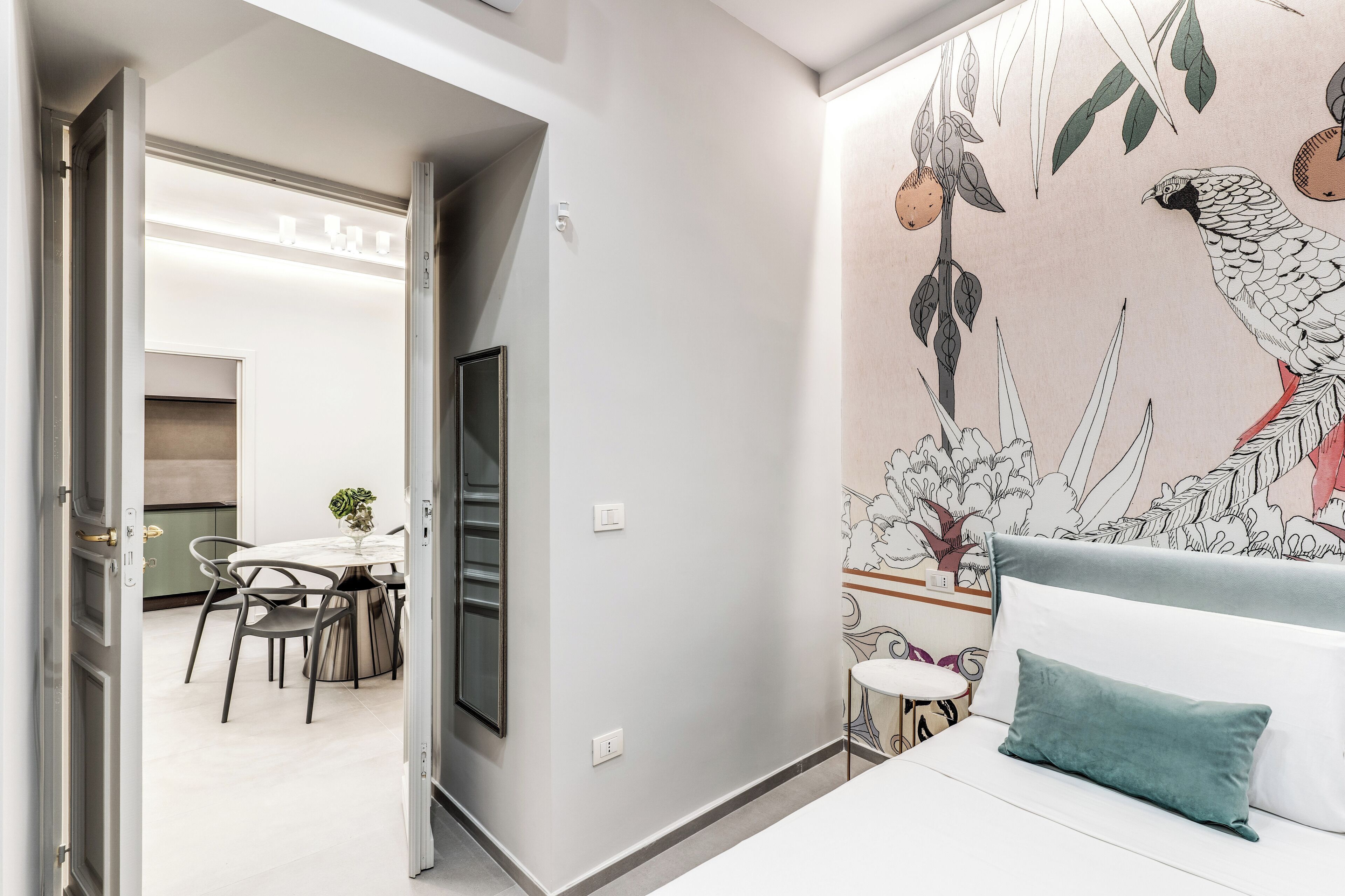 A modern bedroom with a white bed, teal pillow, and a wall mural of birds and foliage.