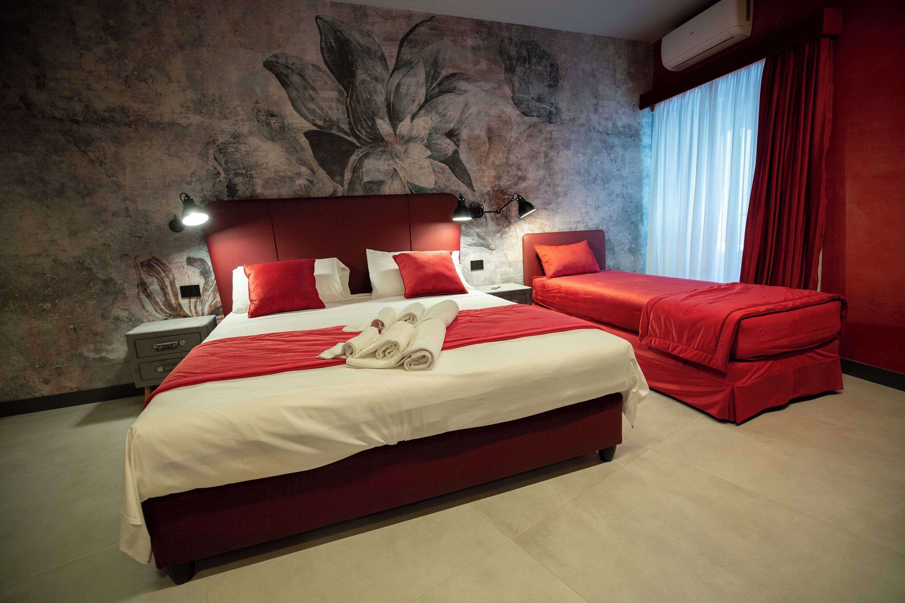 A hotel room with a large bed, a smaller bed, red bedding, a wall mural, and a window with curtains.