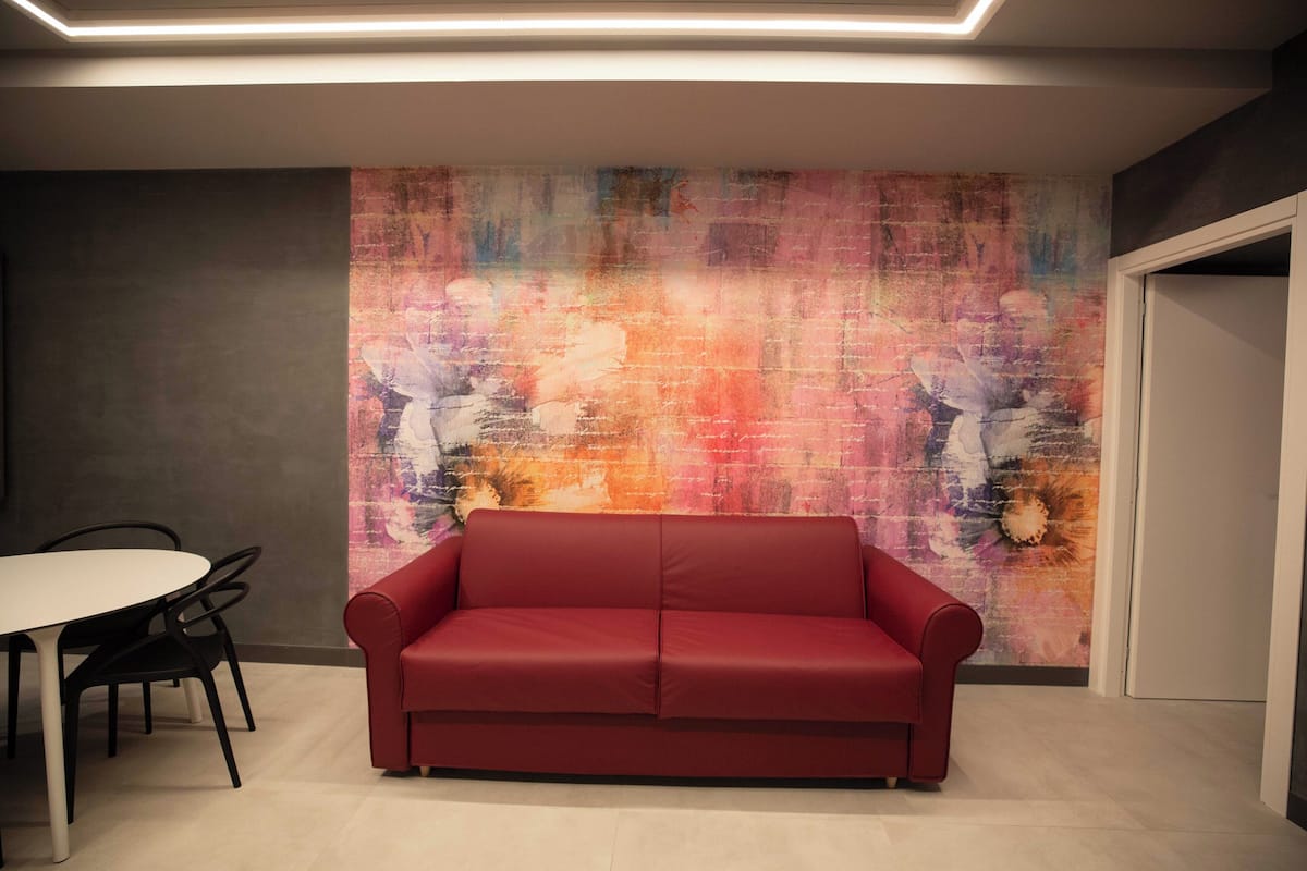 Une pièce moderne avec un canapé rouge, une table de salle à manger ronde entourée de chaises et un mur décoré d’une œuvre abstraite aux couleurs vives.
