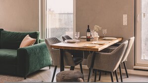 Un comedor con una mesa de madera dispuesta para cuatro personas, rodeada de sillas tapizadas en gris. Se ve un sofá verde con un cojín marrón a la izquierda.
