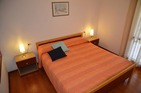 A bedroom with a wooden bed, two bedside tables with lamps, a nightstand, and a framed picture on the wall. - Charming three-room house in a peaceful area (Bibione)