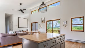 A modern kitchen with a central island, wooden flooring, and large windows offering views of greenery. - Chic Home w/ Fireplace & Beautiful Sunsets (Callicoon)