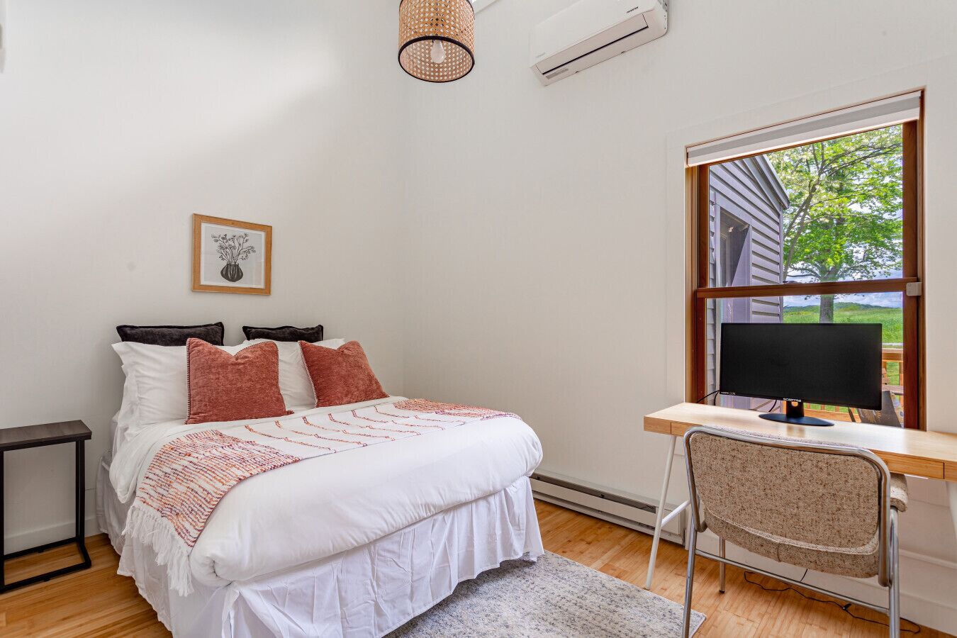 A bedroom with a bed, a desk with a computer, a window with a view of greenery, and an air conditioning unit.