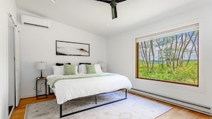 A bedroom with a bed, bedside tables, an air conditioning unit, a ceiling fan, a window with a view of trees, and a painting on the wall. - Chic Home w/ Fireplace & Beautiful Sunsets (Callicoon)