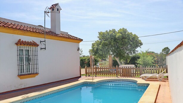 A swimming pool with a white building and a wooden fence in the background. - Casa Ana           . (Conil de la Frontera)