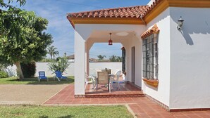 A covered patio with outdoor furniture, a white building with a tiled roof, and a tree with red flowers. - Casa Ana           . (Conil de la Frontera)