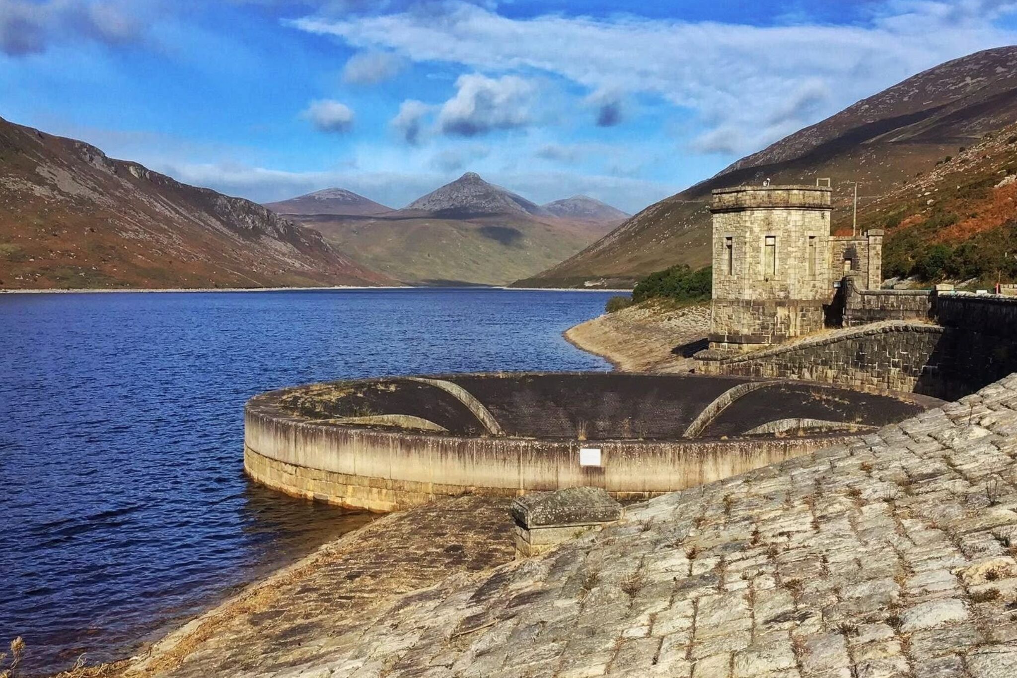Un muro de piedra con una pequeña torre, un lago tranquilo y montañas al fondo.