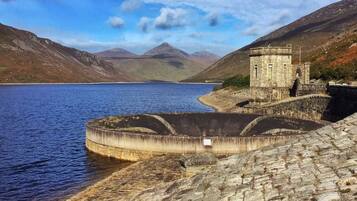 A stone dam with a small tower, a calm lake, and mountains in the background.