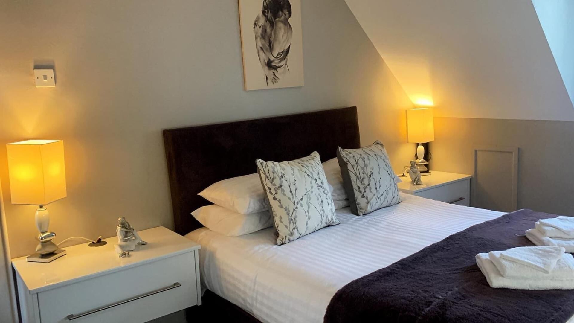 A hotel room with a large bed, two bedside tables with lamps, a headboard, and a painting on the wall.