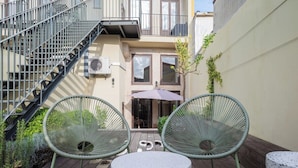 A small outdoor patio with two green wire chairs and a round table, a staircase, and a building with windows and a door in the background.