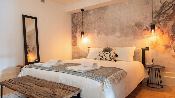 A modern bedroom with a large bed, two pendant lights, a small round table, and a bench.