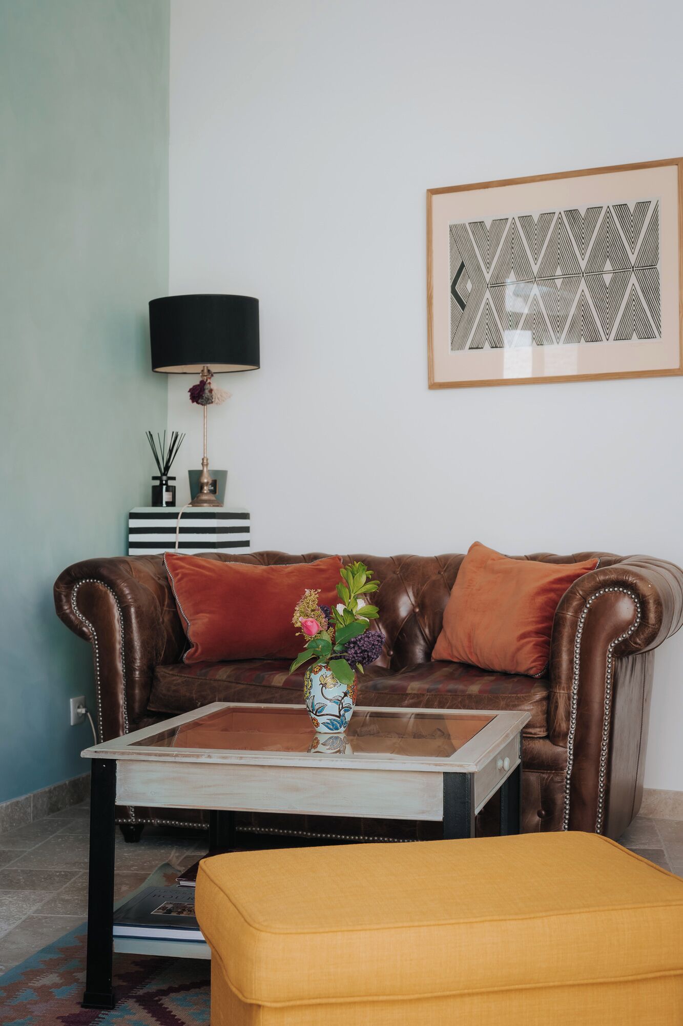 A living room with a brown leather sofa, a coffee table, and a yellow ottoman. There is a framed artwork on the wall and a floor lamp beside the sofa.
