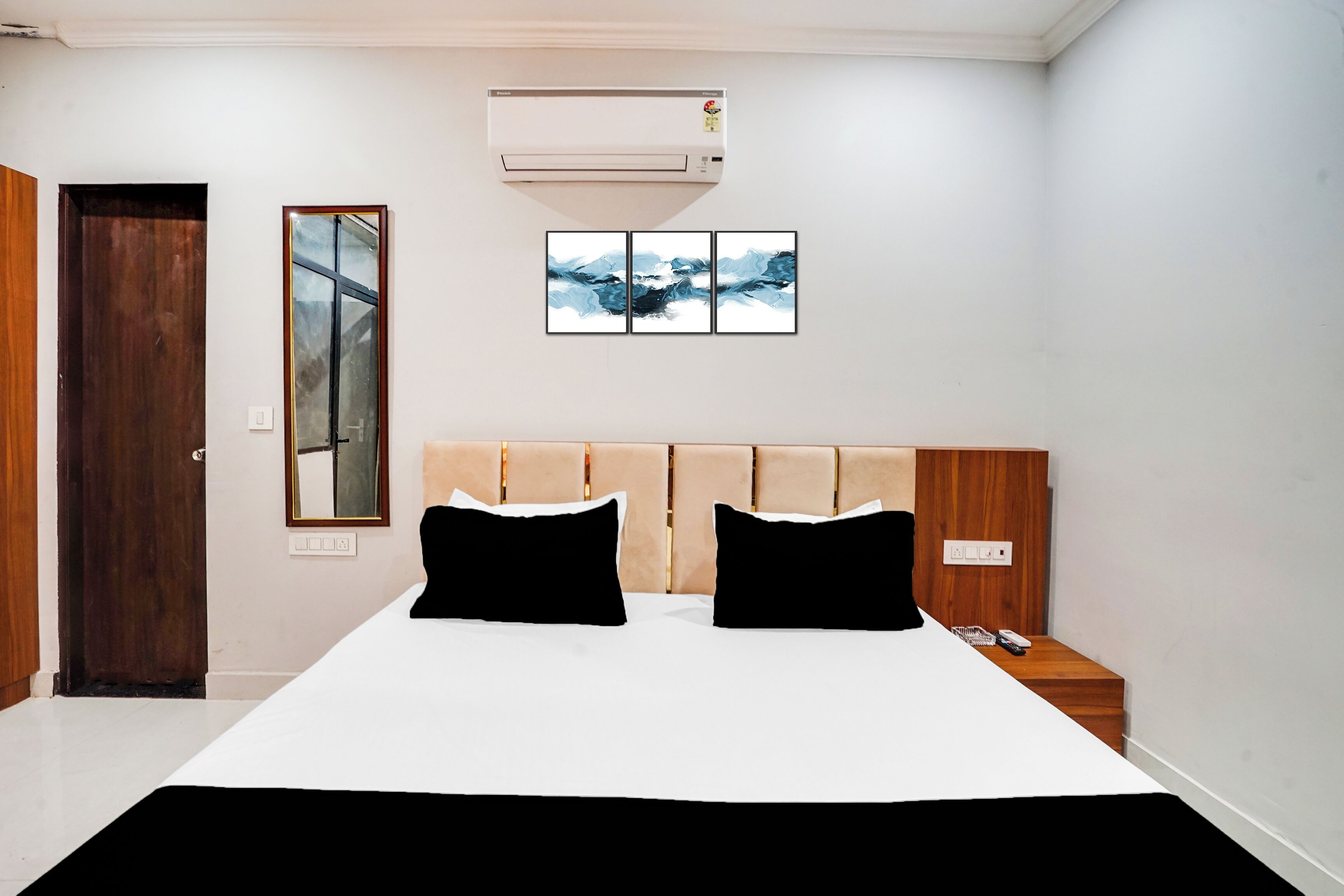 A bedroom with a bed, two black pillows, a wooden headboard, a nightstand, a phone, a wall-mounted air conditioner, a mirror, and three framed artworks.