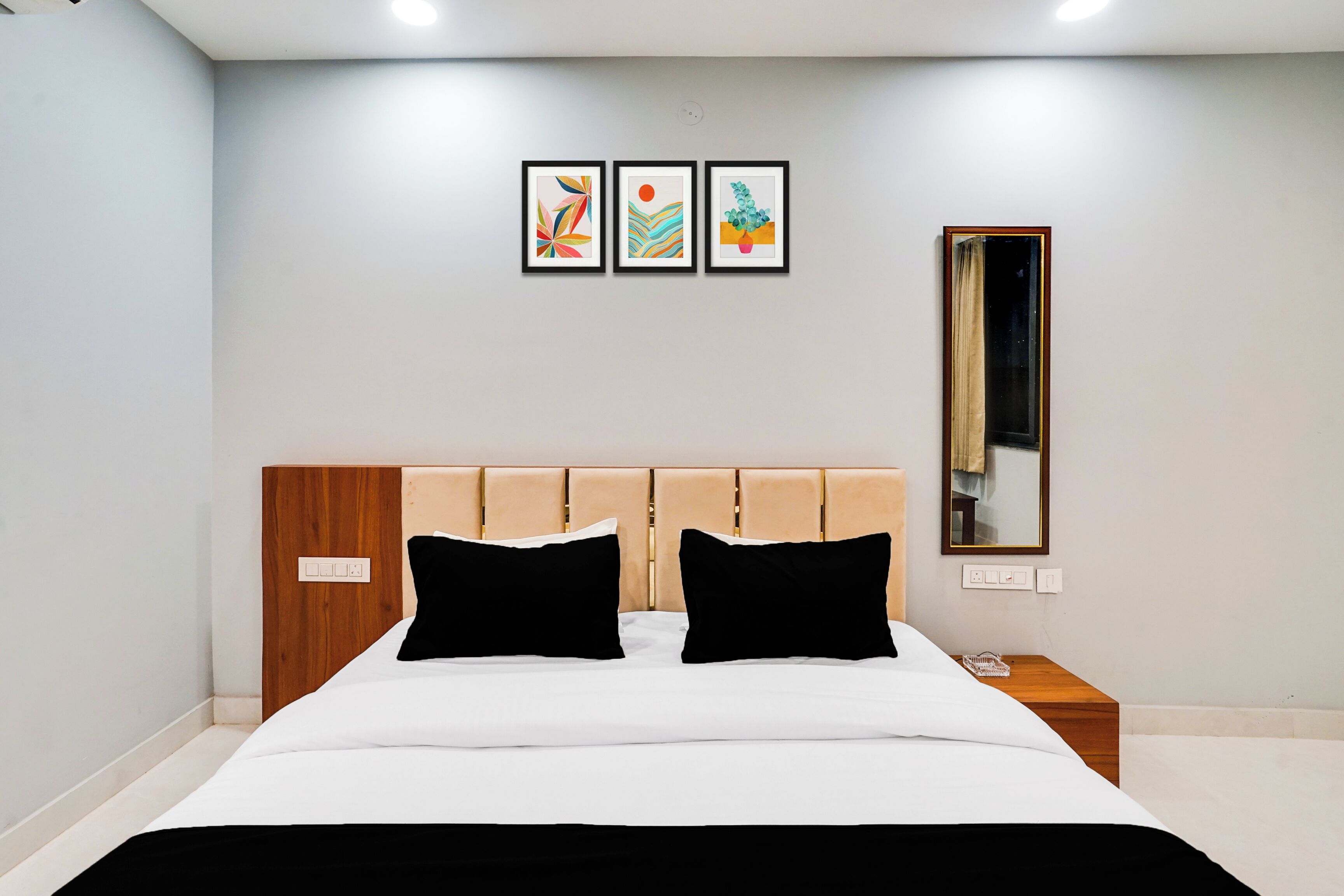 Habitación de hotel moderna con cabecera de madera, una cama grande con ropa de cama blanco y negro, una mesita de noche con lámpara, y tres obras de arte enmarcadas en la pared.