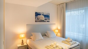 Una habitación de hotel con cama, dos lámparas de noche, una mesita, un cuadro en la pared y un ventanal con cortinas.