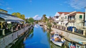 A narrow canal flanked by buildings with balconies and outdoor seating, suggesting a riverside or lakeside location. - Cozy studio with Sea View on the beach in Corfu (94X3+83V Kavos, Greece)