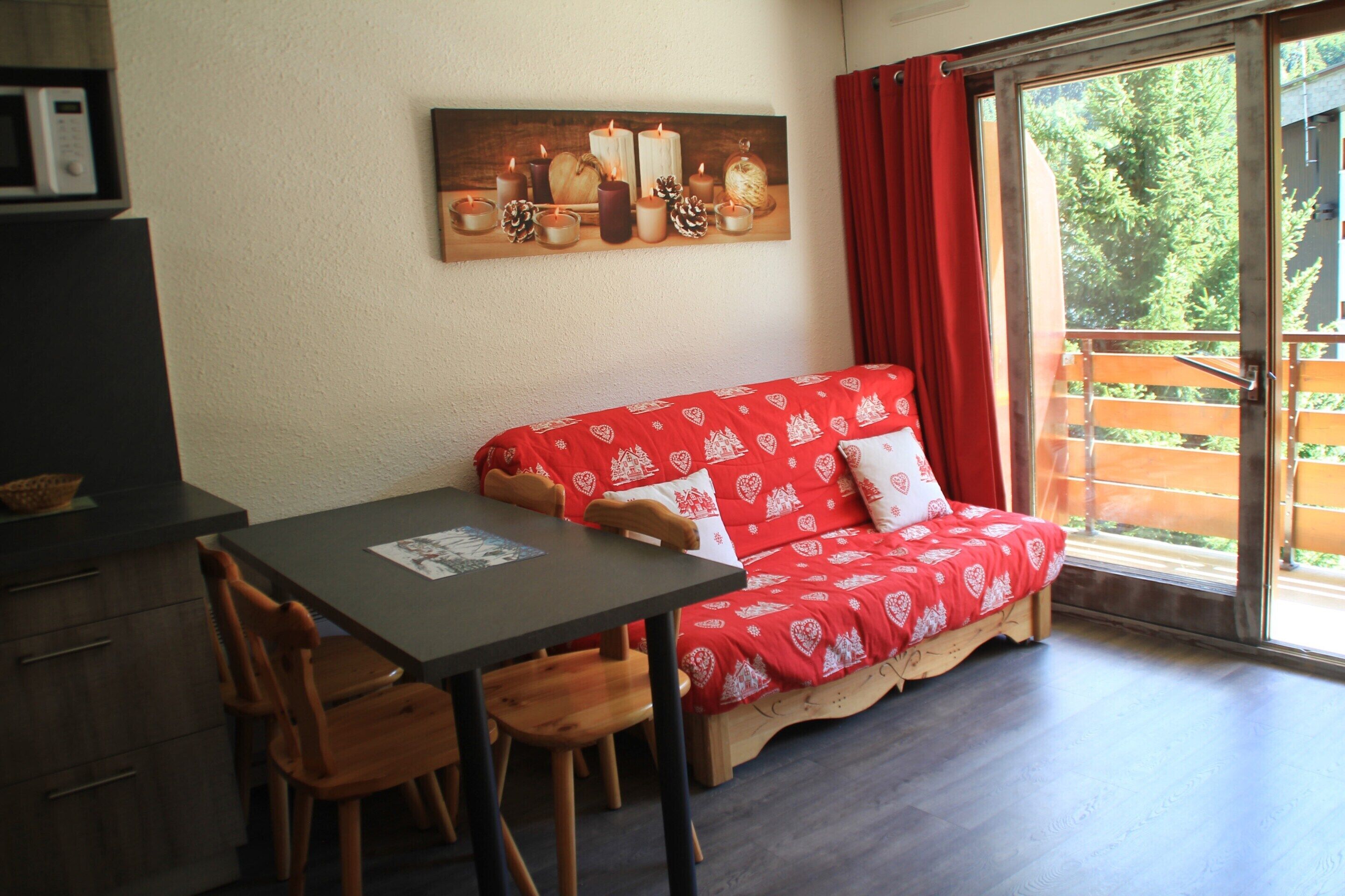 Ein gemütliches Zimmer mit einer roten Couch, einem Holzespeisetisch und Stühlen. Ein Bild von Kerzen und Tannenzapfen hängt an der Wand.