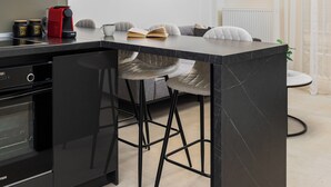 A modern kitchen with a black marble island, white cabinetry, and a built-in oven. There is a coffee maker on the counter and a plant in the corner. - Solange by halu! (Thessaloniki)