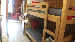 Ein Etagenbett mit grauem Topper und roten Kissen, ein Holzstuhl und ein kleiner Tisch mit einer Lampe.
