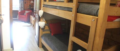 A bunk bed with a grey mattress and red pillows, a wooden chair, and a small table with a lamp.