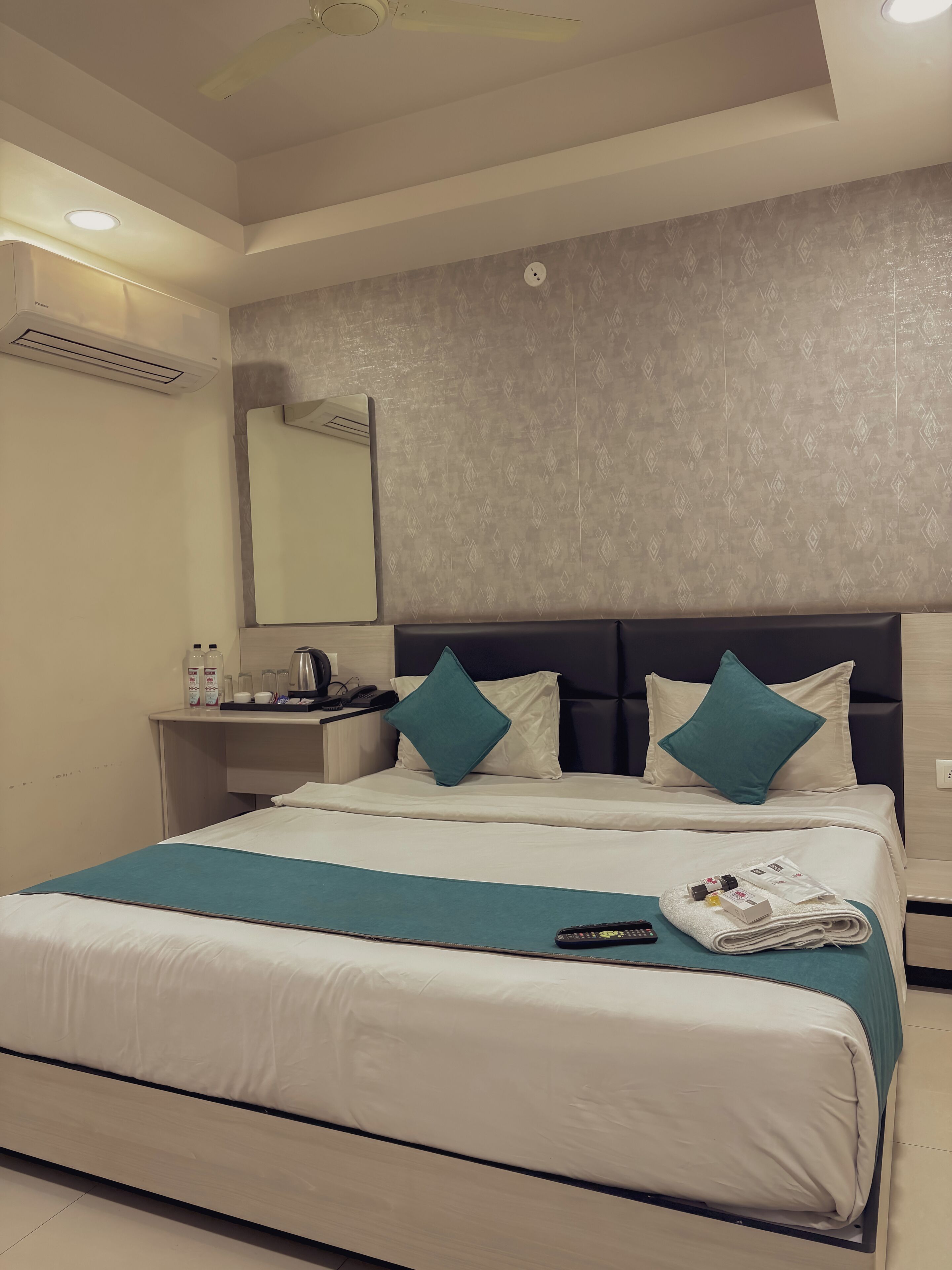 A modern hotel room with a large bed, a bedside table with toiletries, a mirror, and a ceiling fan.