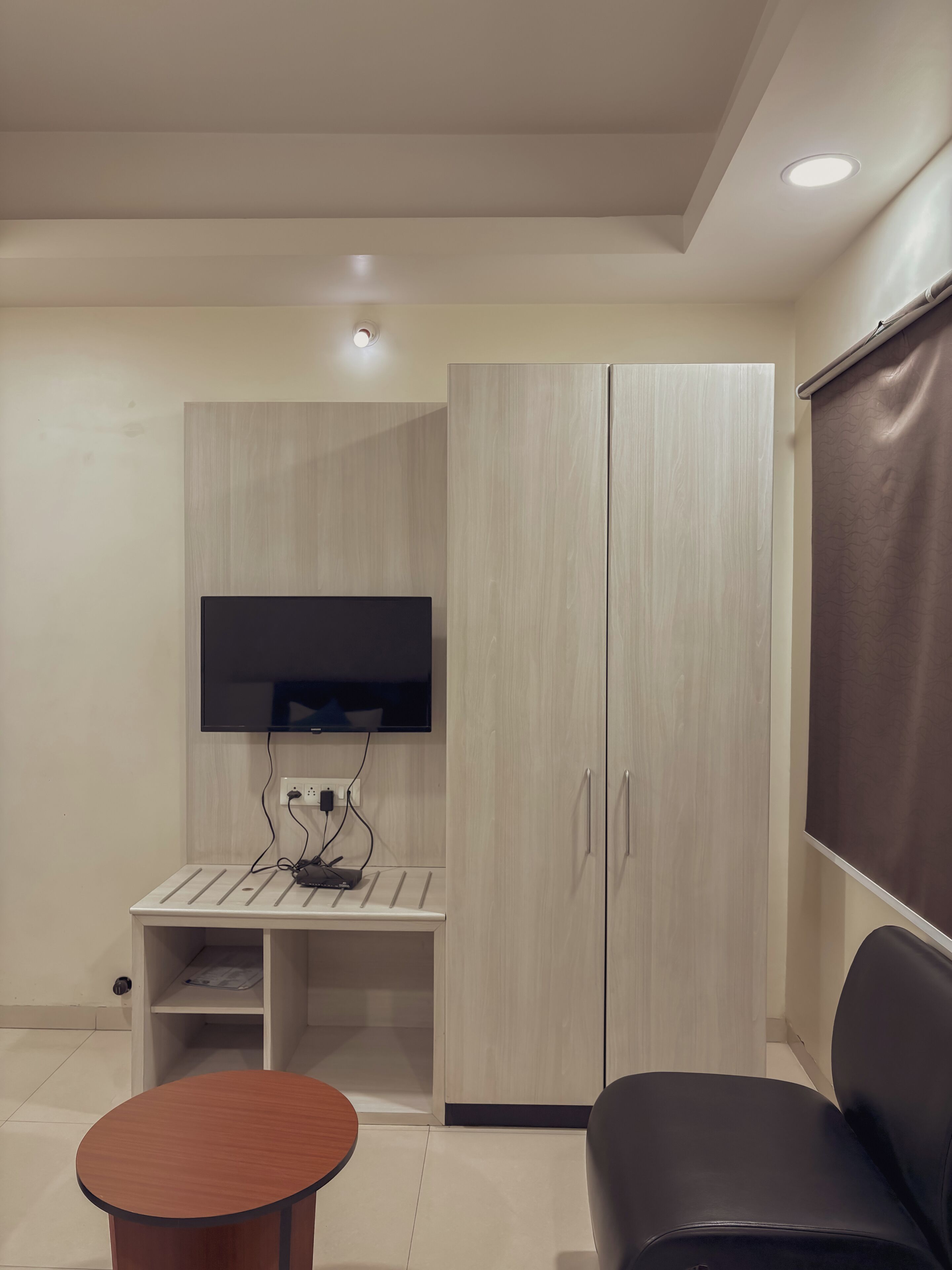 A modern room with a flat-screen TV mounted on a wall, a wooden wardrobe, a small table, and a black sofa.