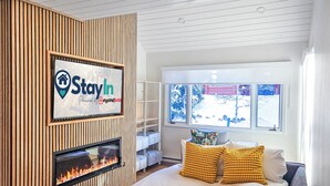Een moderne slaapkamer met een open haard, een bed met gele kussens, een wandgemonteerde televisie en een raam met uitzicht.