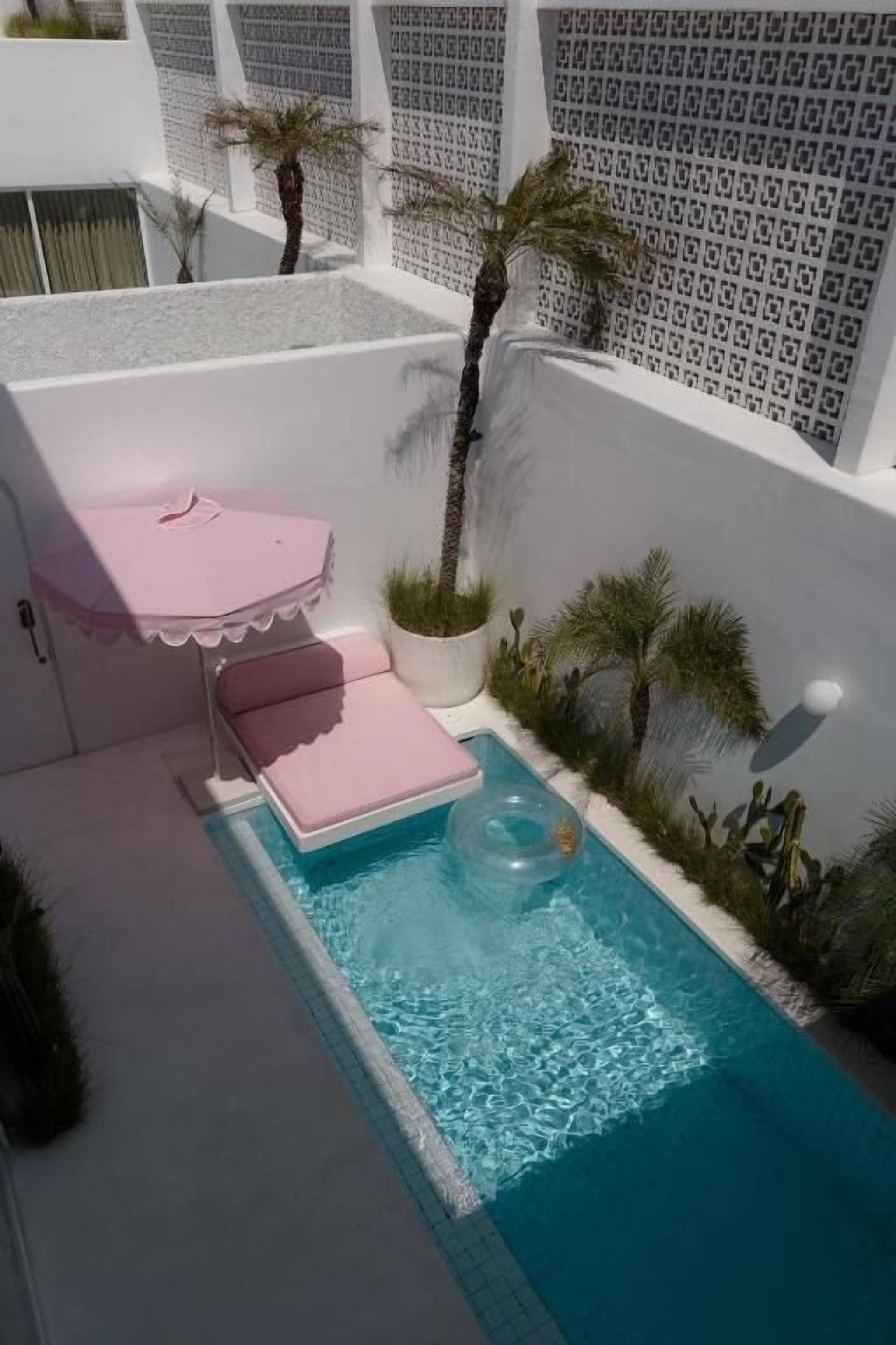 A small pool with a pink lounge chair and umbrella, surrounded by potted plants and a patterned wall.