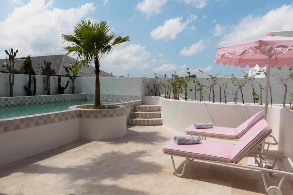 A pool area with two lounge chairs, a pink umbrella, and a palm tree.