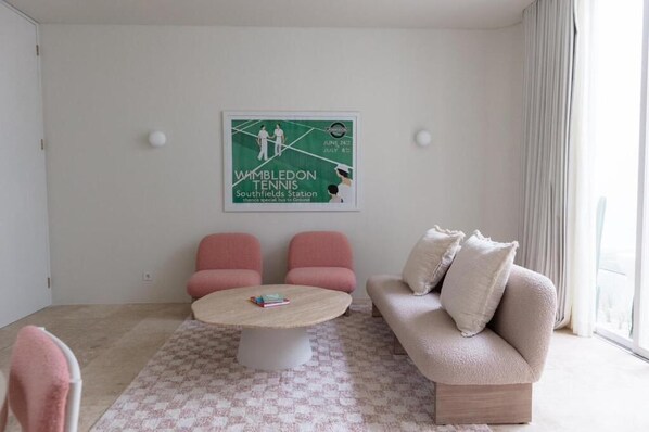 A modern living room with a round coffee table, two pink armchairs, a beige sofa, and a framed Wimbledon poster on the wall.