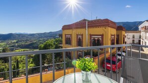 A balcony with a view of a yellow building, a red car, and a mountainous landscape.
