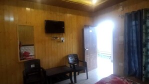 A wooden-paneled room with a bed, a desk, two chairs, a television mounted on the wall, and a door leading to a balcony. - Pine Palace Homestay (Joshimath)